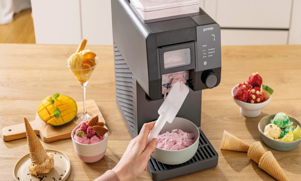 Na drewnianym blacie stoi maszynka do lodów z logo \'gorenje\', z której do miski nakładane są różowe lody. Obok znajdują się różne desery lodowe i owoce, w tym mango i truskawki.