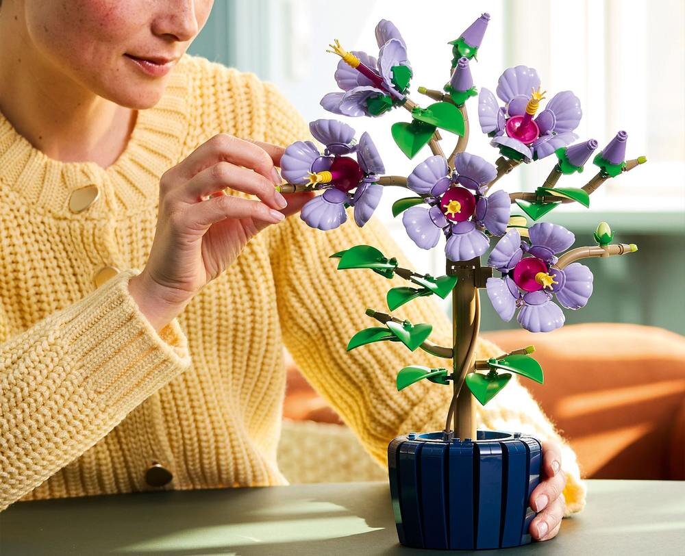 Osoba w żółtym swetrze układa model rośliny z klocków LEGO, przedstawiający kwiaty hibiskusa w fioletowym kolorze. Model umieszczony jest w niebieskiej doniczce.