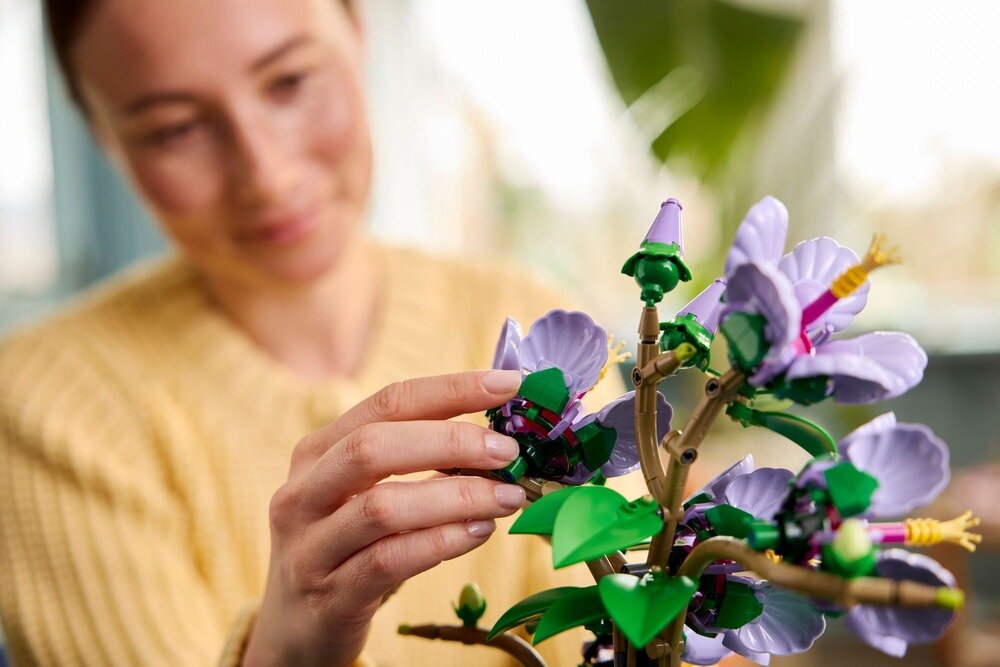 Osoba w żółtym swetrze układa model rośliny z klocków LEGO, przedstawiający kwiaty hibiskusa w odcieniach fioletu i zieleni. W tle widoczne są rozmyte elementy wnętrza.