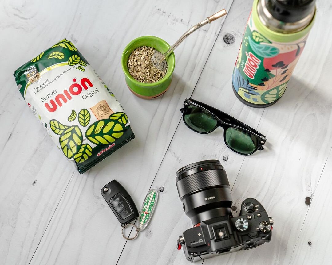 Na drewnianym stole znajduje się opakowanie Yerba Mate Union Suave, zielona tykwa z bombillą, termos z kolorowym wzorem, okulary przeciwsłoneczne, aparat fotograficzny oraz kluczyki z brelokiem. Opakowanie yerby ma zielono-białą grafikę z napisem \'suave unión Original\'.
