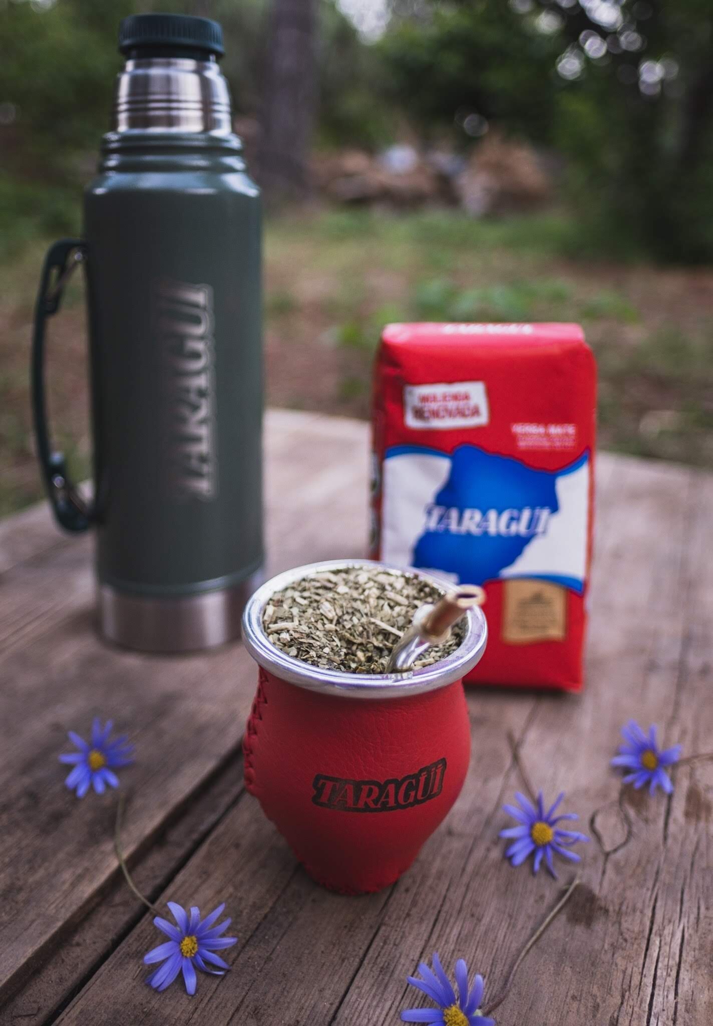 Na drewnianym stole znajduje się termos, paczka Yerba Mate Taragui oraz naczynie z yerbą i bombillą. W tle widoczna jest zieleń, a na stole leżą niebieskie kwiaty.