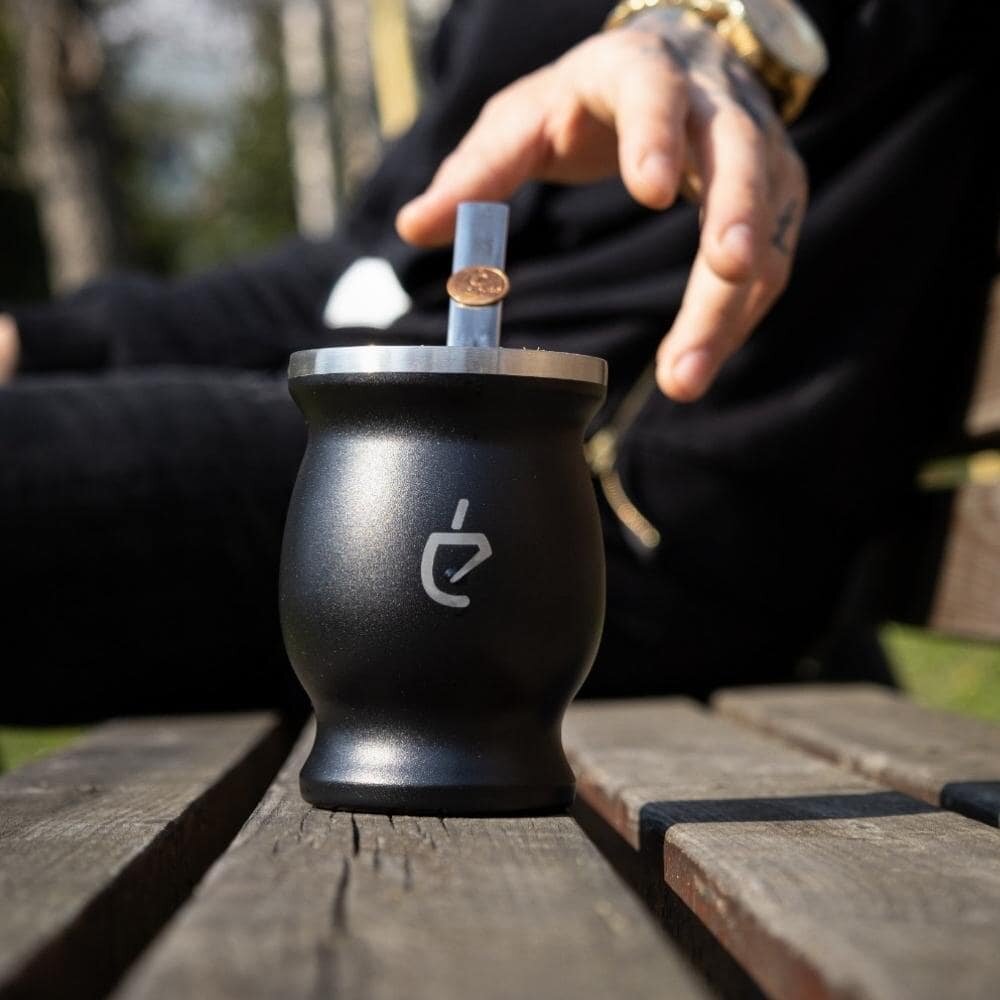 Czarna tykwa do yerba mate z metalowym obrzeżem i logo w kształcie stylizowanego listka. W tle widoczna jest ręka z zegarkiem i tatuażem, sięgająca po tykwę.