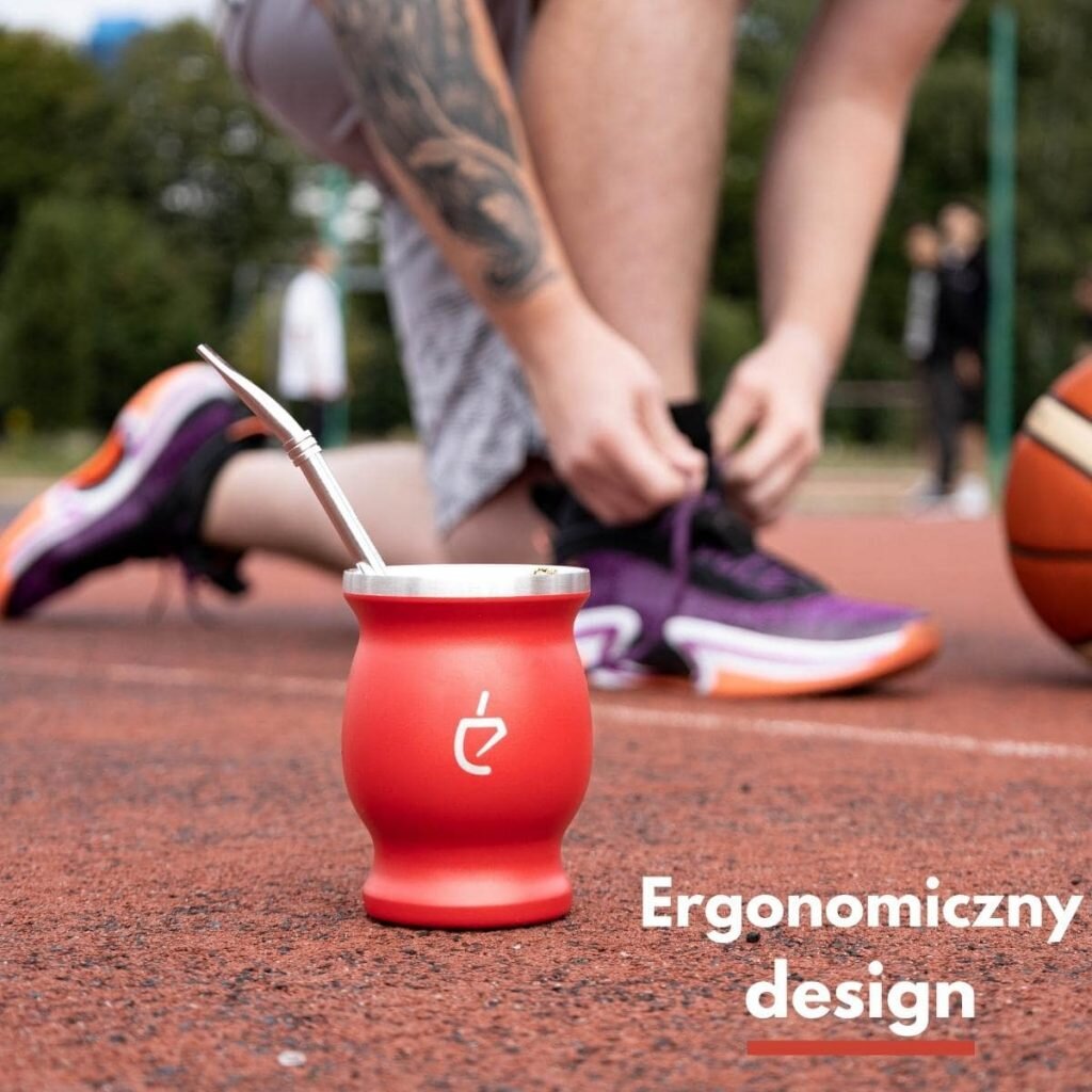 Czerwona tykwa z metalową bombillą stoi na boisku sportowym, w tle osoba wiąże buty sportowe. Na obrazie widoczny jest napis \'Ergonomiczny design\'.