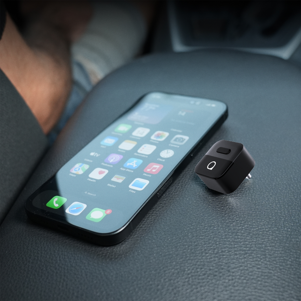 Na ciemnej powierzchni leży smartfon z widocznymi ikonami aplikacji na ekranie. Obok znajduje się małe, czarne urządzenie z przyciskiem i symbolem \'O\'.