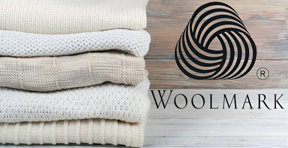 Stos złożonych swetrów w różnych odcieniach beżu i bieli obok logo Woolmark na drewnianym tle.