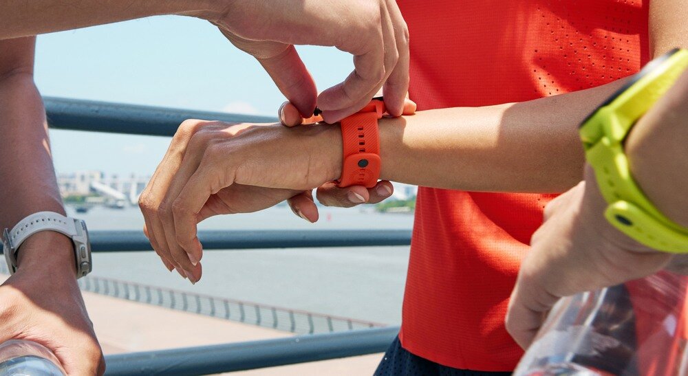 Osoba nosi pomarańczowy smartwatch na nadgarstku, a inna osoba pomaga go zapiąć. W tle widać barierkę i fragment krajobrazu z wodą.