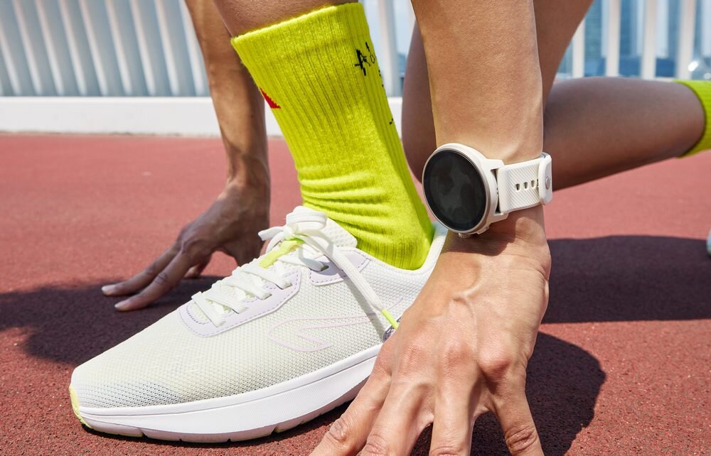 Osoba w jasnych butach sportowych i jaskrawożółtych skarpetkach klęczy na bieżni, na nadgarstku ma biały smartwatch. W tle widoczna jest barierka.