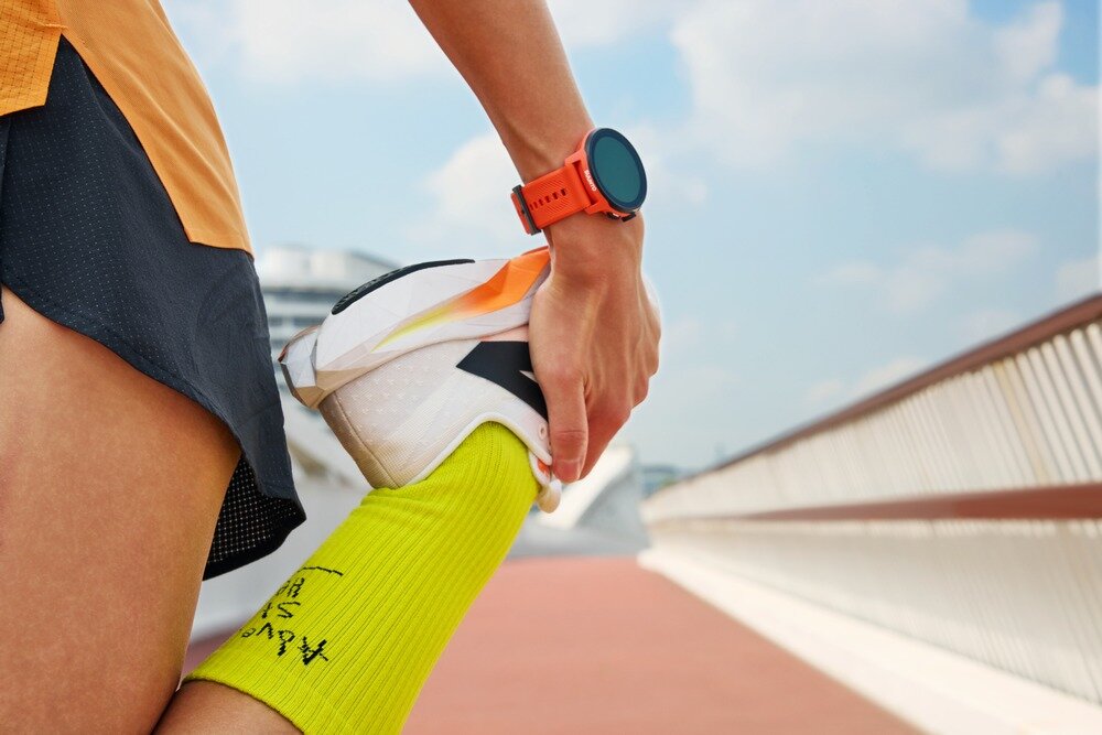 Osoba w sportowym stroju rozciąga nogę, nosząc pomarańczowy smartwatch na nadgarstku. Na skarpetce widoczny jest czarny napis w języku azjatyckim.