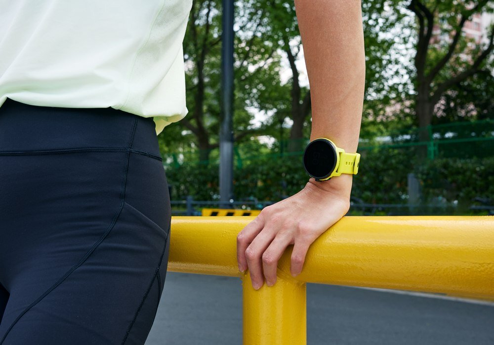 Osoba opiera się o żółtą barierkę, nosząc limonkowy smartwatch na nadgarstku. W tle widoczne są drzewa i fragment ulicy.