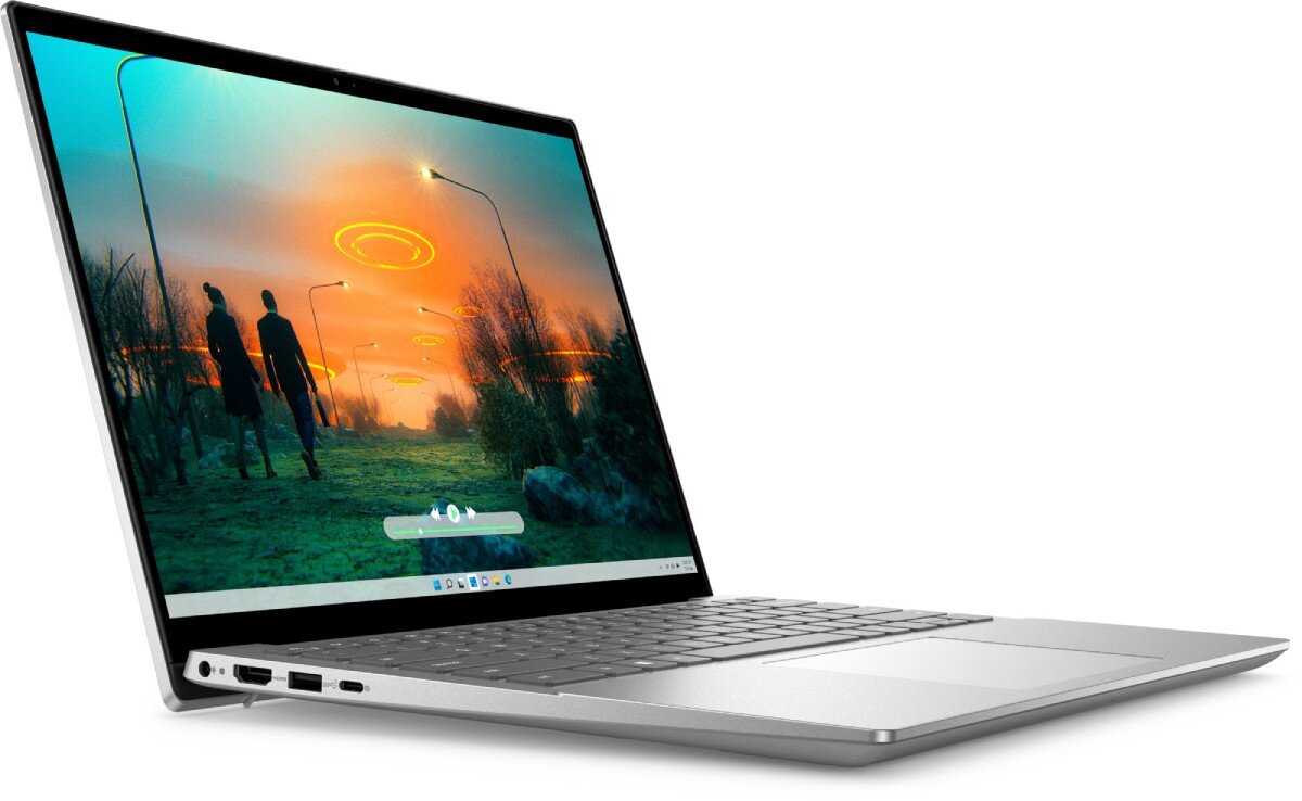 Laptop z otwartym ekranem, na którym wyświetlany jest krajobraz z dwoma postaciami i latarniami ulicznymi. Obudowa jest srebrna, a klawiatura ma ciemne klawisze.