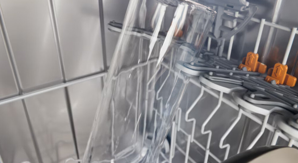 Wnętrze zmywarki z widocznymi szklanymi naczyniami umieszczonymi na metalowych półkach. W tle widoczne są elementy plastikowe w kolorze pomarańczowym.