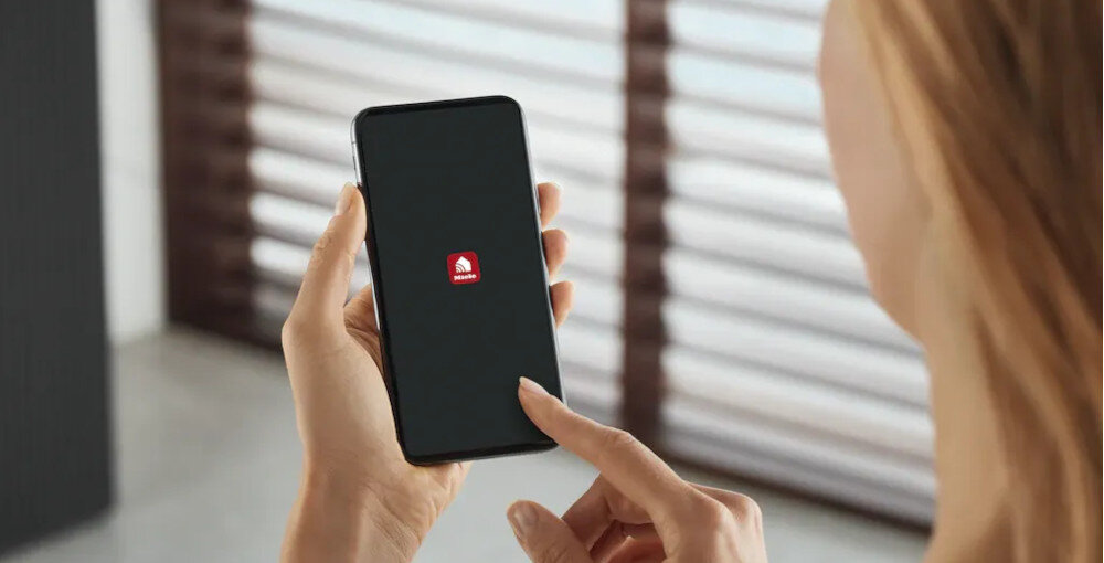 Osoba trzyma smartfon z wyświetlonym logo aplikacji na ekranie. W tle widoczne są rozmyte żaluzje.