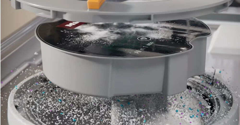 Wnętrze zmywarki z widocznym okrągłym pojemnikiem na detergent, na którym znajduje się proszek. Na pojemniku widoczny jest napis \'Miele\'.