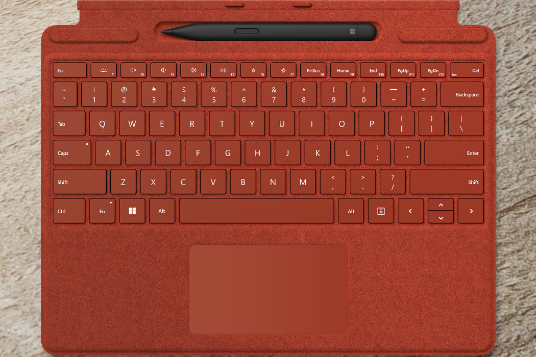 Czerwona klawiatura z pełnym układem klawiszy, w tym klawisze funkcyjne i strzałki, oraz touchpad poniżej. Na górze znajduje się pióro cyfrowe w specjalnym uchwycie.