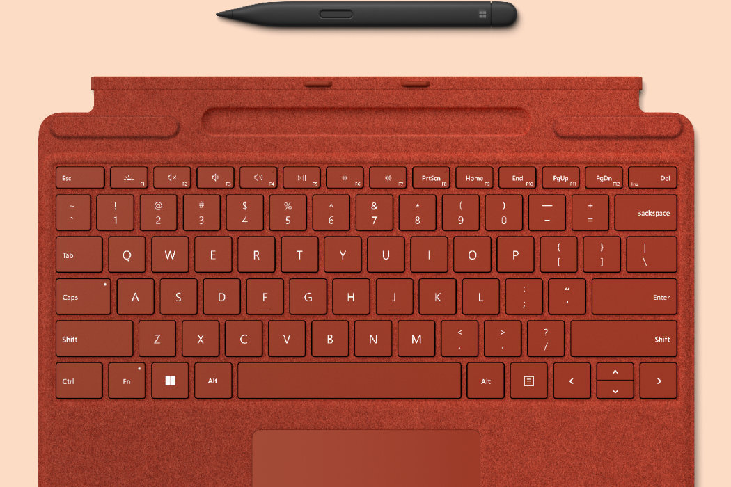 Czerwona klawiatura z układem QWERTY, z widocznymi klawiszami funkcyjnymi i strzałkami. Nad klawiaturą znajduje się smukłe pióro w kolorze czarnym.