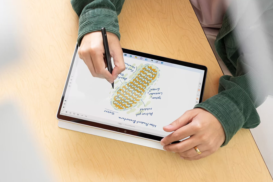 Osoba używa pióra do rysowania na tablecie, na którym widoczny jest kolorowy schemat z notatkami. Tablet leży na drewnianym stole.