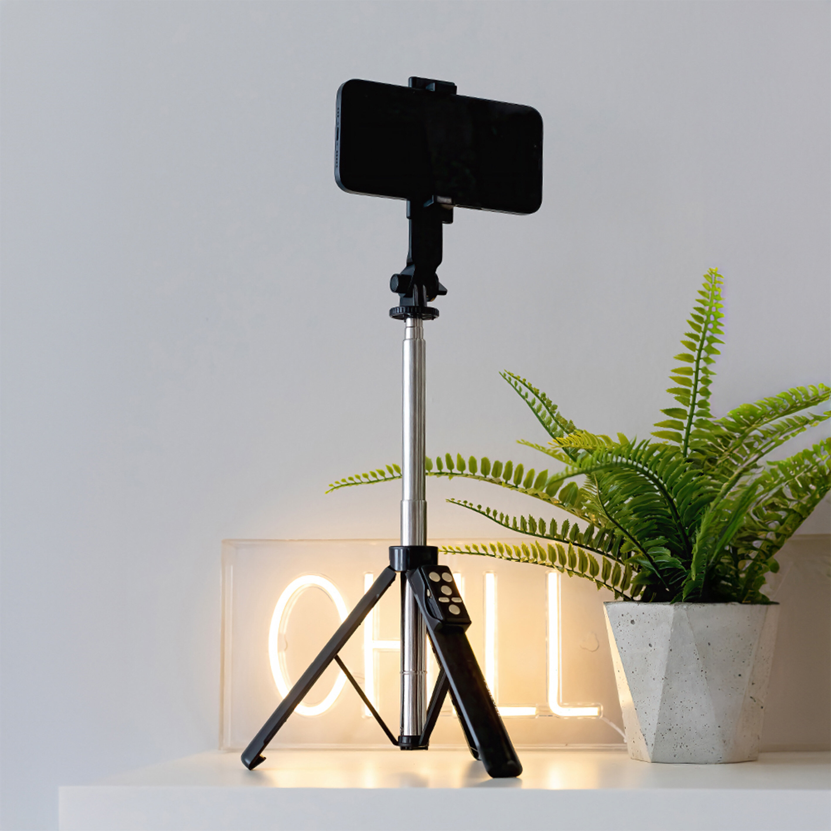 Na białej powierzchni stoi czarny selfie stick z funkcją statywu, na którym zamocowany jest smartfon. Obok znajduje się zielona roślina w doniczce, a w tle widoczny jest napis \'CHILL\'.