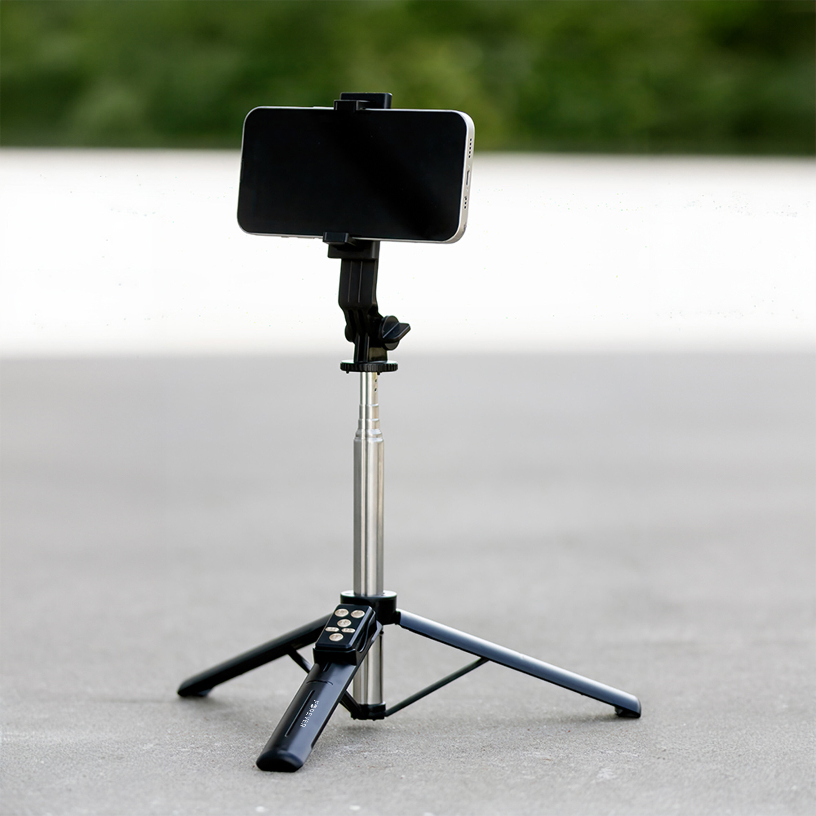 Na zdjęciu widać czarny selfie stick z funkcją statywu, na którym zamocowany jest smartfon. Urządzenie stoi na szarej powierzchni, a w tle widoczna jest rozmyta zieleń.