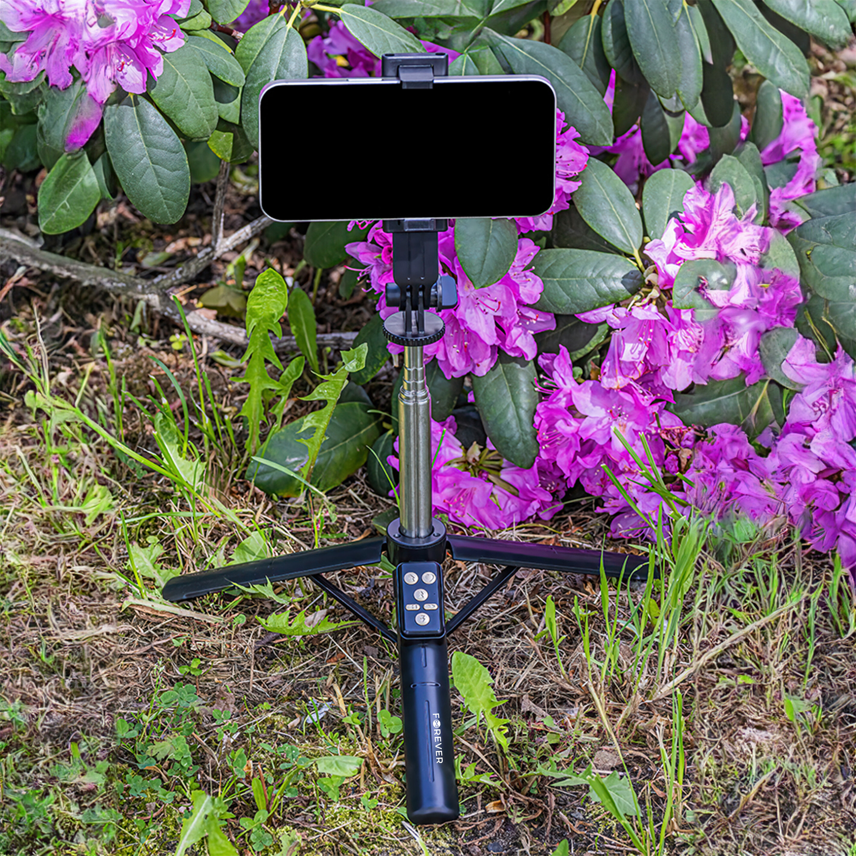 Czarny selfie stick z funkcją statywu stoi na trawie przed różowymi kwiatami. Na uchwycie widoczny jest napis \'FOREVER\'.