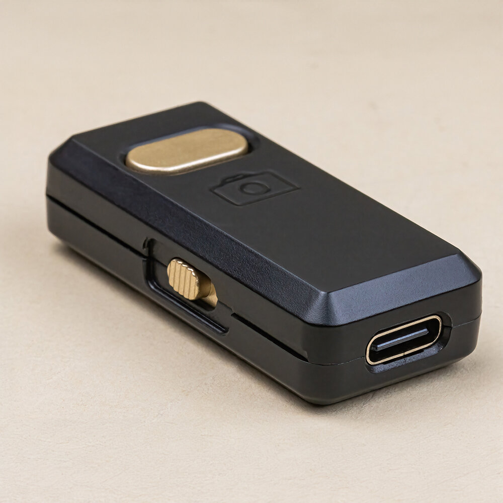 Czarny prostokątny element z metalicznym przyciskiem i symbolem aparatu na górze. Posiada port USB-C i suwak na boku.