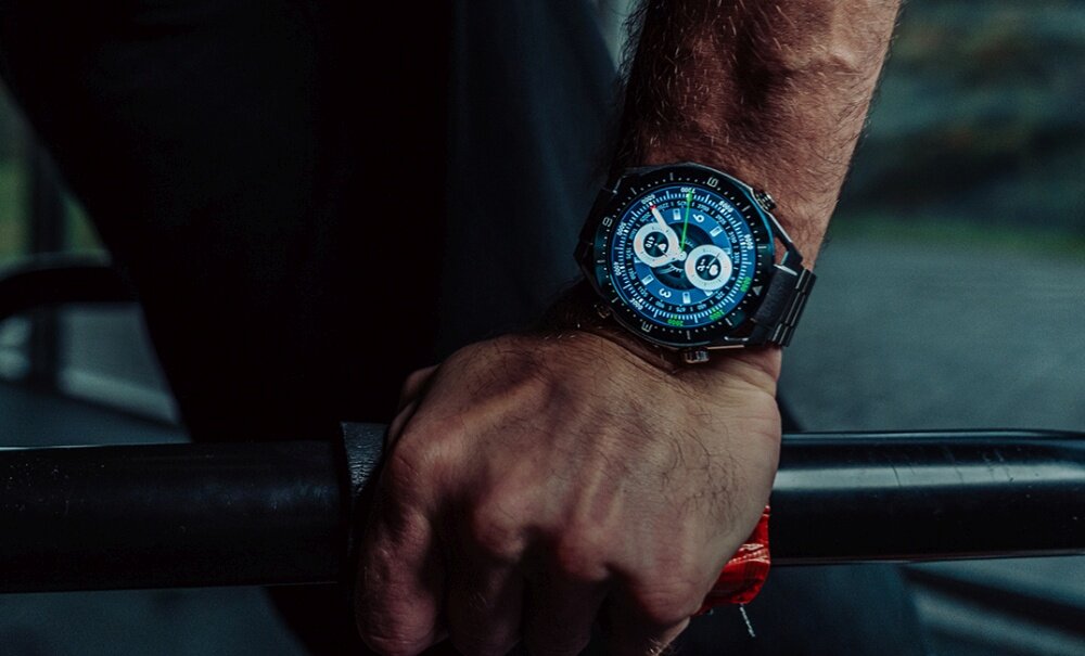 Osoba trzymająca czarny smartwatch z okrągłym, kolorowym wyświetlaczem na nadgarstku, oparta o metalowy pręt.