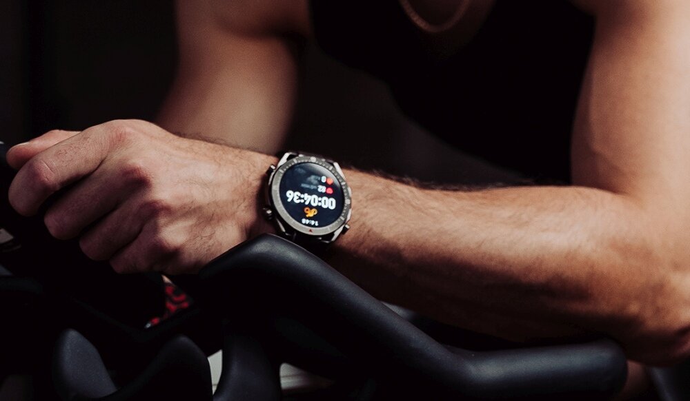 Osoba trzyma kierownicę roweru, na nadgarstku ma czarny smartwatch z wyświetlaczem pokazującym czas 00:00:36 i ikonę serca.