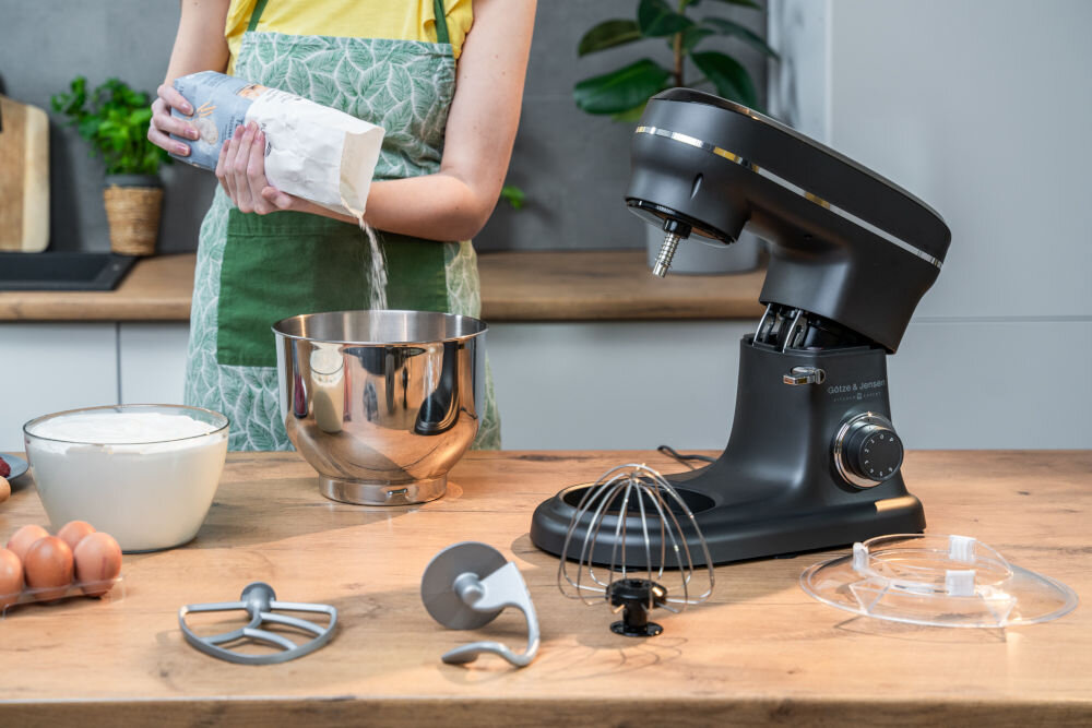 Na blacie kuchennym znajduje się czarny robot kuchenny planetarny GÖTZE & JENSEN SB400K z różnymi końcówkami do mieszania. Obok robota osoba wsypuje mąkę do metalowej miski, a na blacie leżą jajka i miska z mąką.