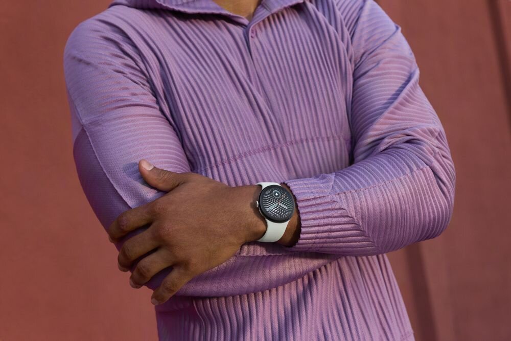 Osoba w fioletowej bluzie z założonymi rękami nosi smartwatch z jasnoszarym paskiem i ciemnym wyświetlaczem.