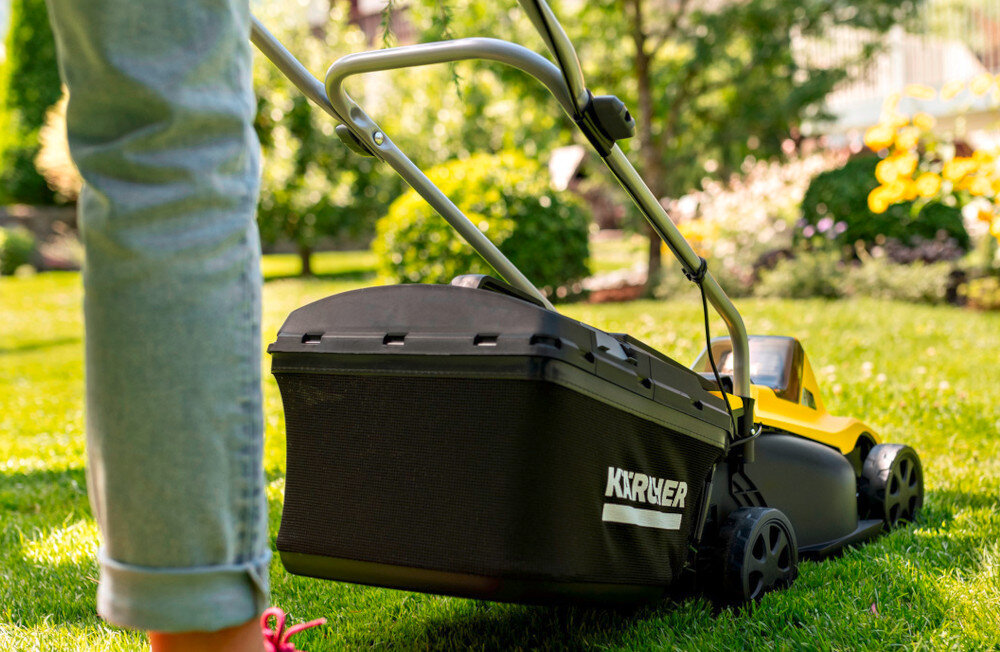 Osoba prowadzi kosiarkę akumulatorową po trawniku w ogrodzie. Na koszu kosiarki widoczne jest logo \'KARCHER\'.