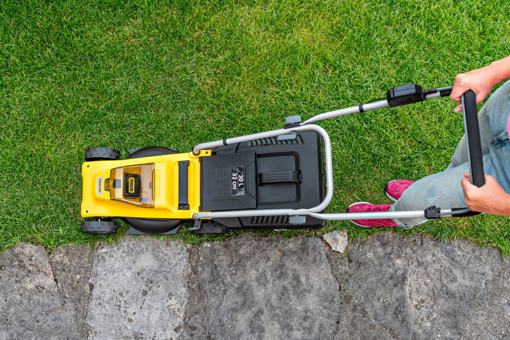 Osoba prowadzi żółtą kosiarkę akumulatorową po trawniku obok kamiennej ścieżki. Na kosiarce widoczne jest logo \'KARCHER\' oraz napis \'30 L 32 cm\'.