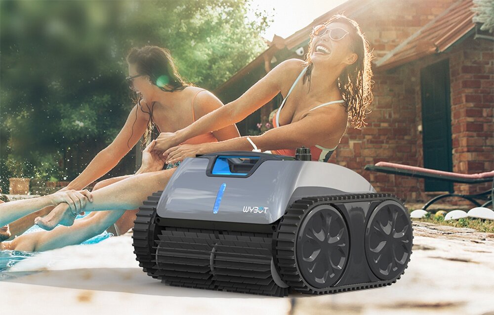Na pierwszym planie znajduje się robot do czyszczenia basenów z widocznym napisem \'WYBOT\'. W tle widać osoby bawiące się przy basenie oraz budynek z cegły.