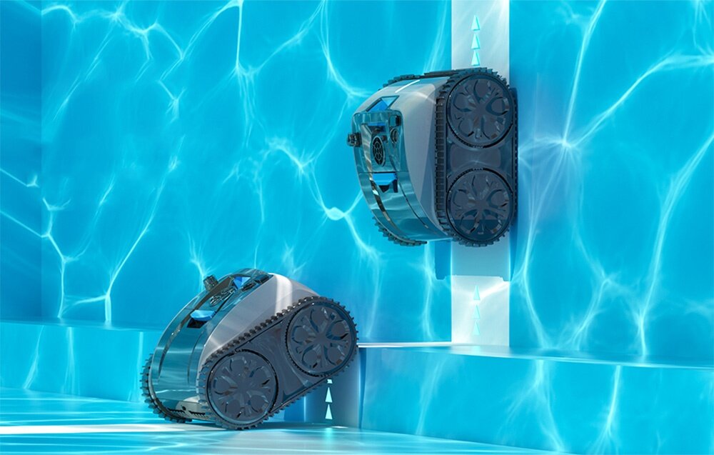 Dwa roboty do czyszczenia basenów poruszają się po ścianach i dnie basenu wypełnionego wodą. Roboty mają gąsienicowe koła i są w kolorze szaro-niebieskim.