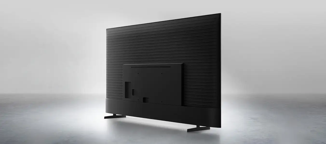 2026-metalstream-design-pc 1352x600 Obraz przedstawia tył czarnego telewizora z widocznymi poziomymi żłobieniami i prostokątnym panelem montażowym. Telewizor stoi na dwóch prostych nóżkach na jasnym tle.