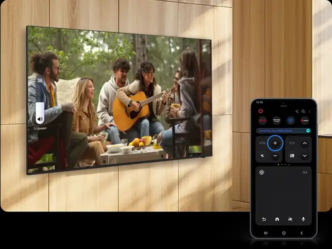 Na ścianie wisi telewizor wyświetlający obraz grupy osób siedzących na zewnątrz, jedna z nich gra na gitarze. Obok znajduje się smartfon z aplikacją do sterowania telewizorem, na ekranie widoczne są przyciski do regulacji głośności i zmiany kanałów.