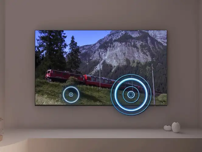 Telewizor wyświetla obraz z pociągiem jadącym przez górski krajobraz, z nałożonymi niebieskimi okręgami symbolizującymi dźwięk. Na półce pod telewizorem znajdują się dwa dekoracyjne elementy.