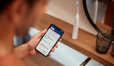 Osoba trzyma smartfon z aplikacją pokazującą statystyki dotyczące szczotkowania zębów. Na blacie obok stoi biała szczoteczka soniczna oraz szklanka.