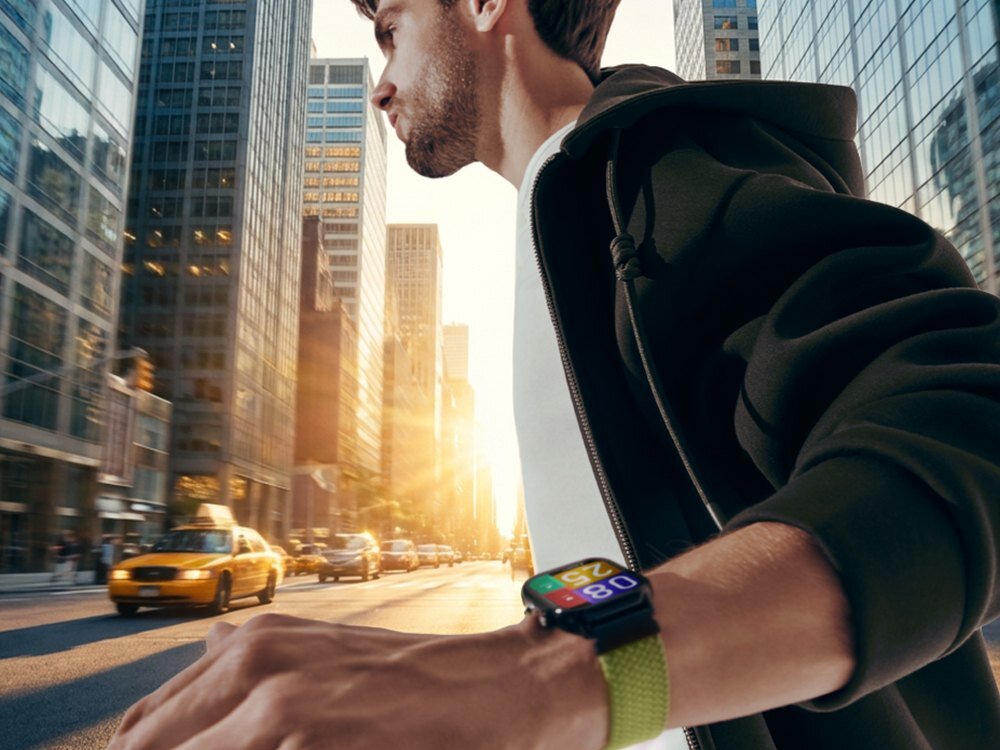 Osoba w ciemnej bluzie biegnie ulicą w mieście, nosząc smartwatch z kolorowym wyświetlaczem na nadgarstku. W tle widać wysokie budynki i przejeżdżające żółte taksówki.