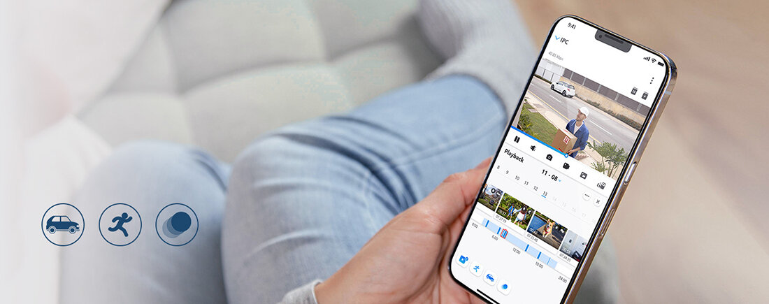 Osoba trzyma smartfon, na którym wyświetlana jest aplikacja monitoringu z widokiem z kamery. Obok widoczne są trzy ikony: samochód, biegnąca postać i księżyc.