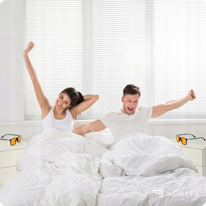 Dwie osoby siedzą na łóżku, przeciągając się, w jasnym pokoju z zasłoniętymi żaluzjami. Na szafkach nocnych znajdują się okulary z pomarańczowymi szkłami, a w rogu widoczne jest logo \'NOBLUE\'.
