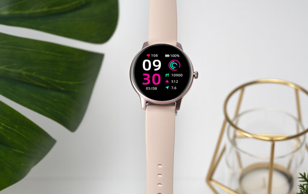 Smartwatch z różowym paskiem i okrągłym wyświetlaczem pokazującym godzinę 09:30, datę 05/08 oraz dane dotyczące zdrowia i aktywności. W tle widoczne są zielone liście i złoty świecznik.