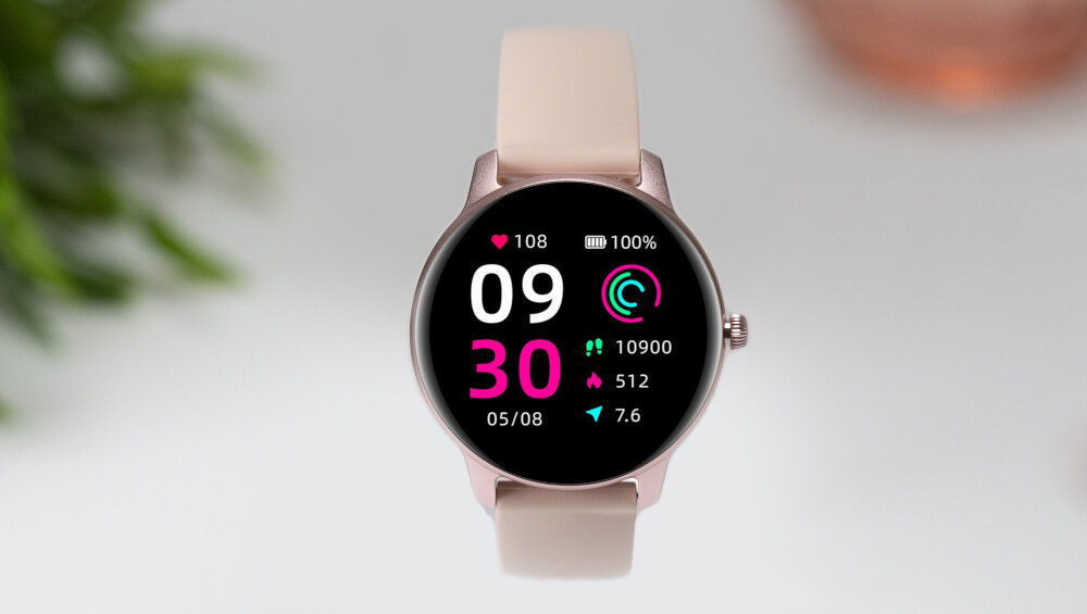 Smartwatch z różowym paskiem i okrągłym wyświetlaczem pokazuje godzinę 09:30, datę 05/08 oraz różne wskaźniki zdrowotne, takie jak tętno 108 i kroki 10900. Tło jest rozmyte, z zielonymi liśćmi po lewej stronie.