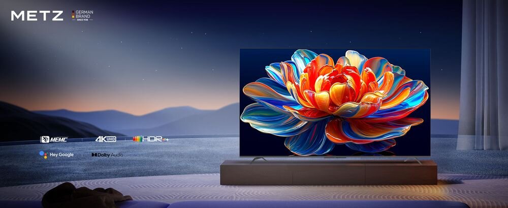 Telewizor wyświetla kolorowy obraz kwiatu na tle ciemnego, górskiego krajobrazu. Widoczne są logotypy: METZ, MEMC, 4K UHD, HDR, Hey Google, Dolby Audio.