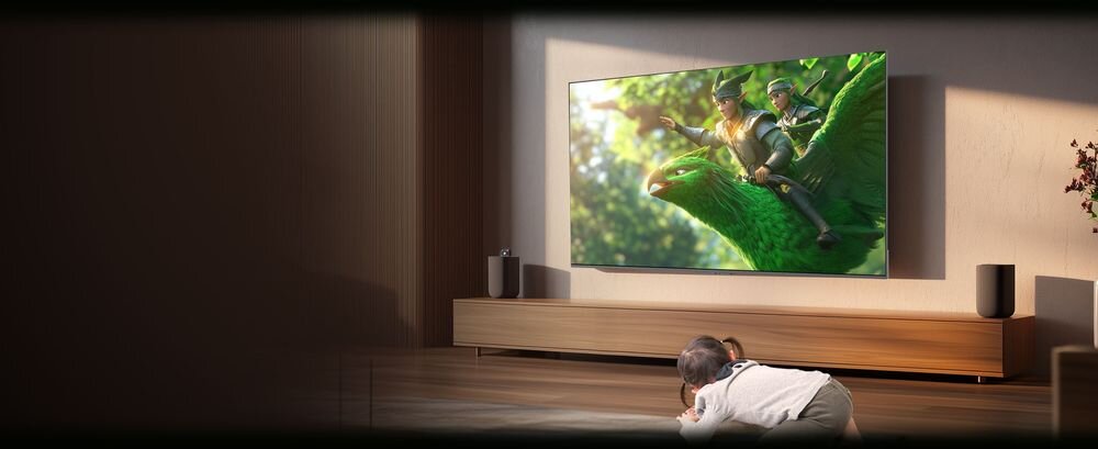Telewizor wyświetla scenę z animacji, na której dwie postacie jadą na dużym zielonym ptaku. Przed telewizorem na podłodze leży dziecko, a obok znajdują się dwa głośniki.