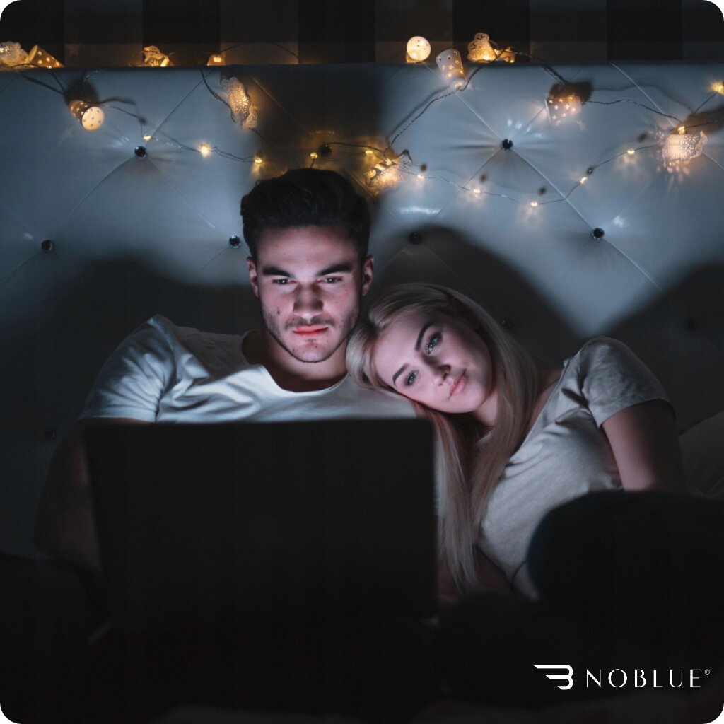 Dwie osoby siedzą na łóżku, patrząc na ekran laptopa. W tle widoczne są światełka dekoracyjne, a w prawym dolnym rogu znajduje się logo \'NOBLUE\'.