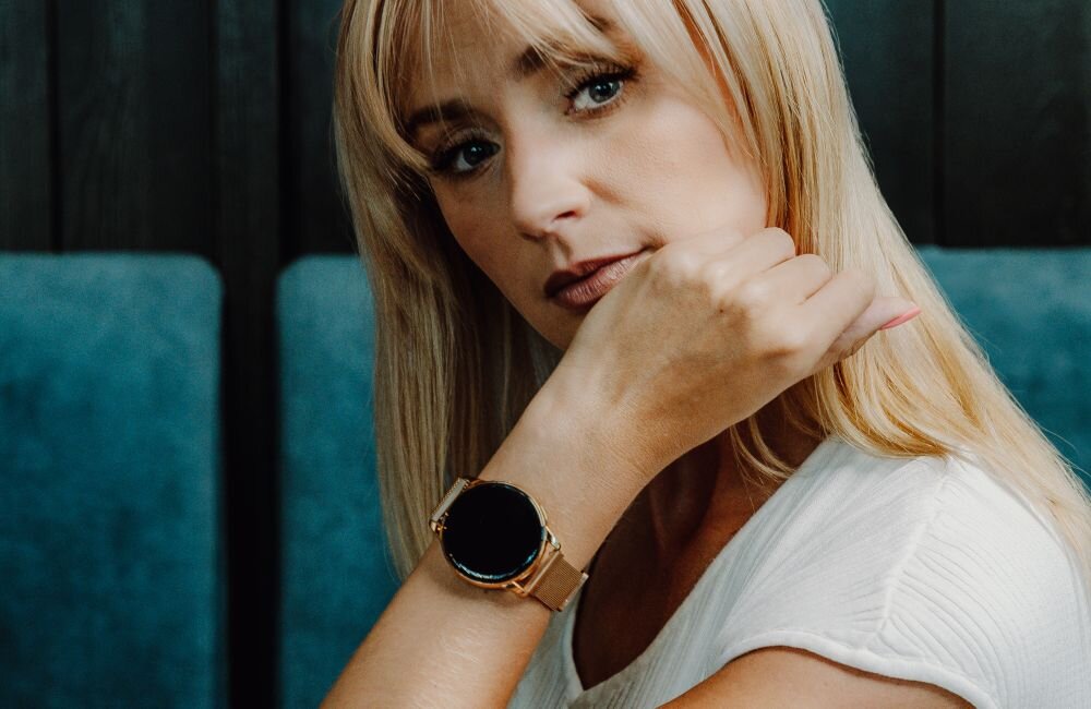 Osoba nosi złoty smartwatch na nadgarstku, siedząc na tle ciemnych paneli i niebieskiego obicia. Smartwatch ma okrągły wyświetlacz i złotą bransoletę.
