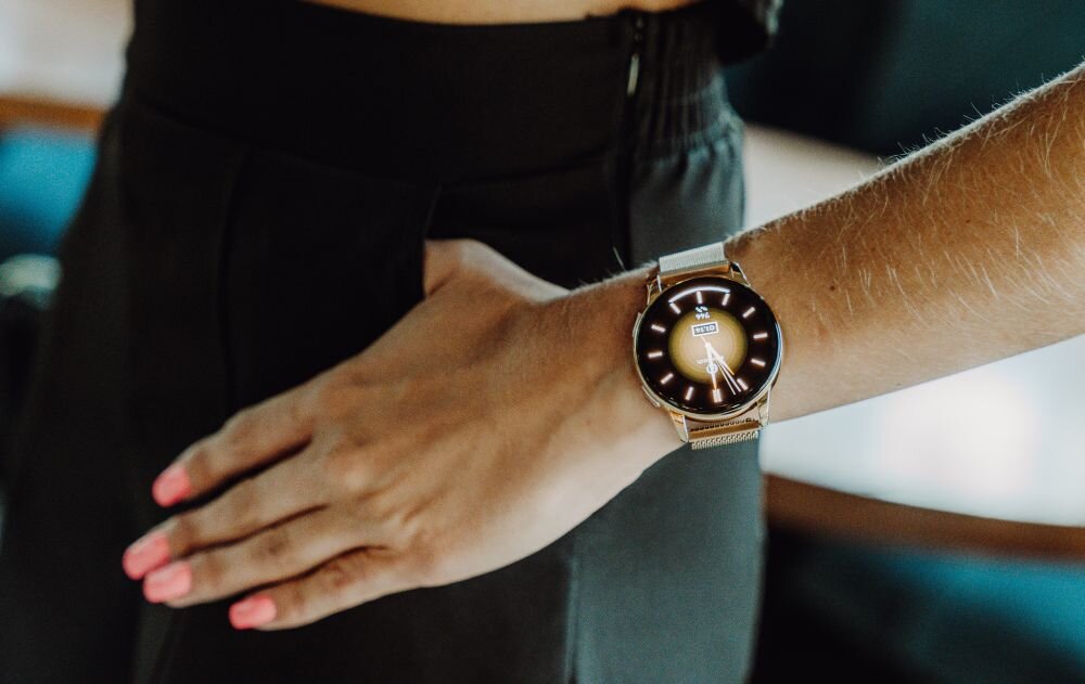 Osoba nosi złoty smartwatch z okrągłą tarczą i wyświetlaczem pokazującym godzinę 10:10. Pasek zegarka jest metalowy i ma siatkową strukturę.