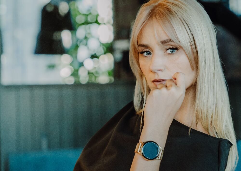 Osoba z długimi blond włosami nosi złoty smartwatch z okrągłym wyświetlaczem. W tle widoczne są rozmyte elementy wnętrza.