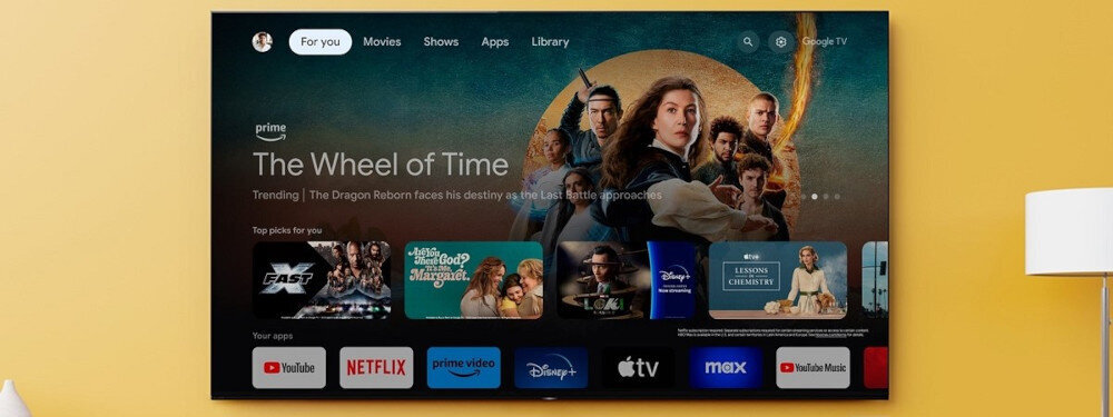Telewizor wyświetla ekran główny z aplikacjami takimi jak YouTube, Netflix, Prime Video, Disney+, Apple TV, Max i YouTube Music. Na ekranie widoczny jest także baner promujący serial \'The Wheel of Time\'.