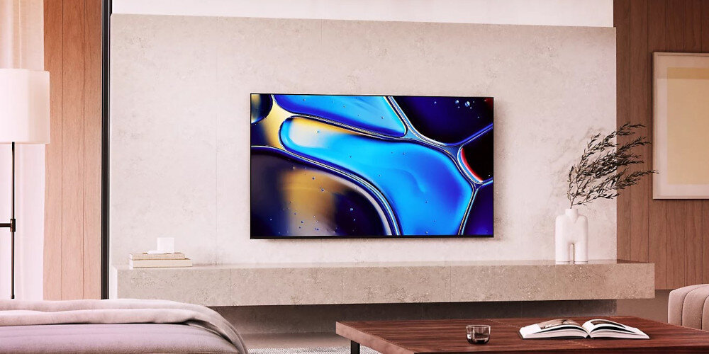 Telewizor zamontowany na ścianie w nowoczesnym salonie, wyświetlający abstrakcyjny obraz w odcieniach niebieskiego i żółtego. W pomieszczeniu znajdują się także stolik z książką i kanapa.