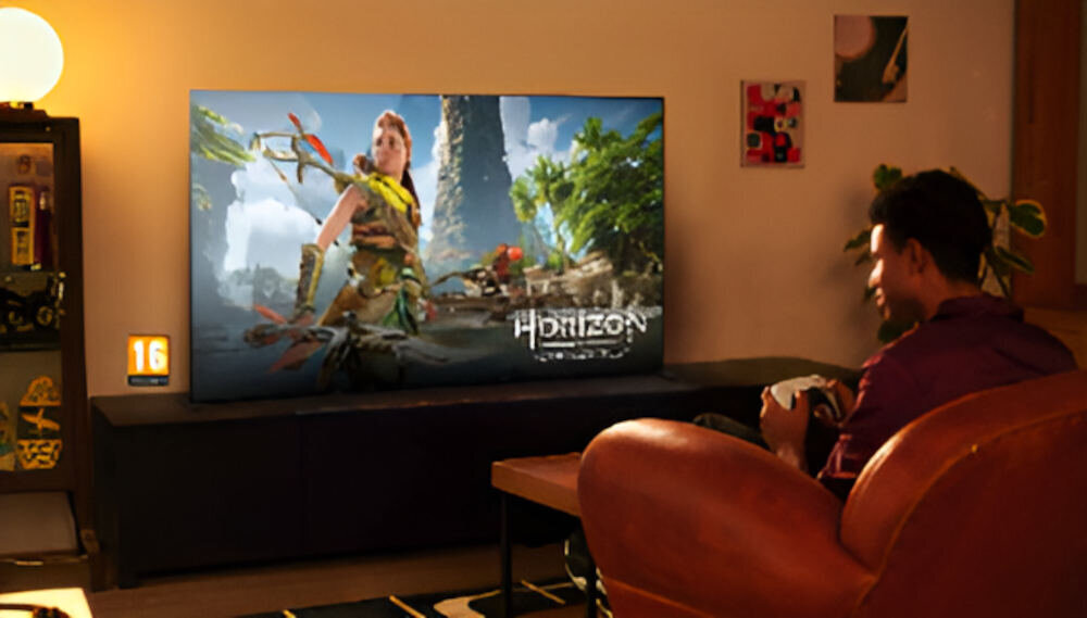 W salonie na szafce stoi duży telewizor wyświetlający grę z postacią w scenerii przyrodniczej. Obok telewizora siedzi osoba trzymająca kontroler do gier.